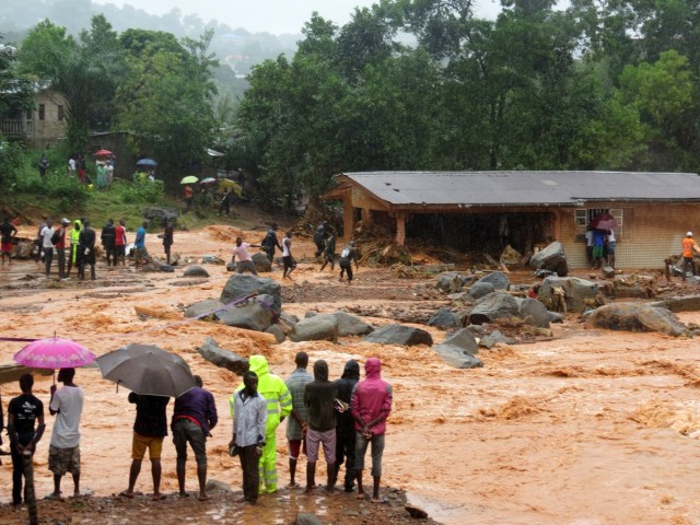 Bystanders look on as floodwaters rage past a damaged building in an area of Freetown on August 14, 2017, after landslides struck the capital of the west African state of Sierra Leone. At least 312 people were killed and more than 2,000 left homeless when heavy flooding hit Sierra Leone's capital of Freetown, leaving morgues overflowing and residents desperately searching for loved ones. An AFP journalist at the scene saw bodies being carried away and houses submerged in two areas of the city, where roads turned into churning rivers of mud and corpses were washed up on the streets. / AFP PHOTO / SAIDU BAH