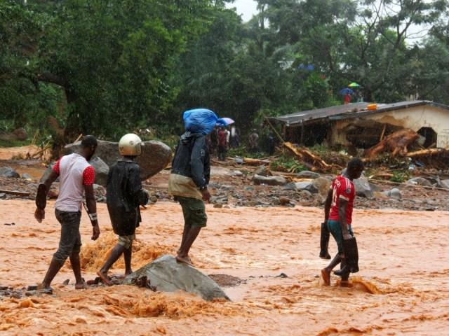 Residents walk through floodwaters past a damaged building in an area of Freetown on August 14, 2017, after landslides struck the capital of the west African state of Sierra Leone.                At least 312 people were killed and more than 2,000 left homeless when heavy flooding hit Sierra Leone's capital of Freetown, leaving morgues overflowing and residents desperately searching for loved ones. An AFP journalist at the scene saw bodies being carried away and houses submerged in two areas of the city, where roads turned into churning rivers of mud and corpses were washed up on the streets.  / AFP PHOTO / SAIDU BAH