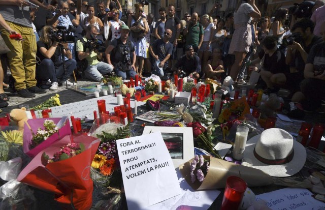 People stand next to flowers, candles, a sign reading ' Out terrorism and attacks, we want peace'  and other items set up on the Las Ramblas boulevard in Barcelona as they pay tribute to the victims of the Barcelona attack, a day after a van ploughed into the crowd, killing 13 persons and injuring over 100 on August 18, 2017. Police hunted for the driver who rammed a van into pedestrians on an avenue crowded with tourists in Barcelona, leaving 13 people dead and  more than 100 injured, just hours before a second assault in a resort along the coast. / AFP PHOTO / PASCAL GUYOT