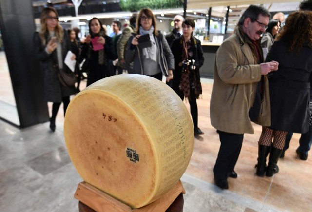 People walk past a loaf of parmesan cheese on display at a stand during a press tour at FICO Eataly World agri-food park in Bologna on November 9, 2017. FICO Eataly World, said to be the world's biggest agri-food park, will open to the public on November 15, 2017. The free entry park, widely described as the Disney World of Italian food, is ten hectares big and will enshrine all the Italian food biodiversity. / AFP PHOTO / Vincenzo PINTO