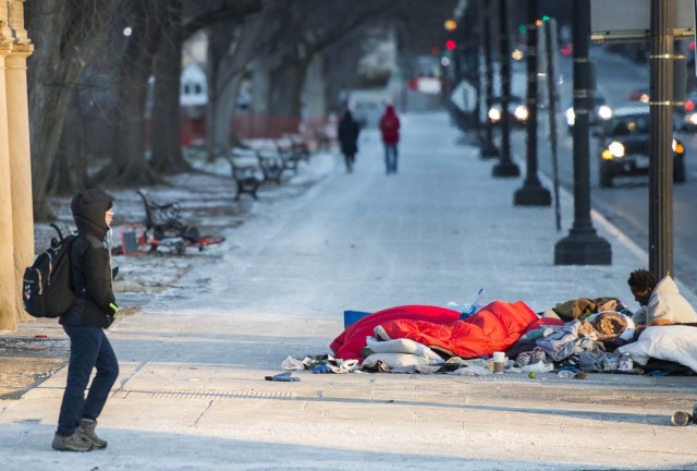 A homeless man rests on top of a subway vent grate as a tourist walks past in Washington, DC on January 5, 2018. The National Weather Service said that very cold temperatures and wind chills will follow for much of the eastern third of the US through the weekend. A cold wave gripping a large section of the United States had already been blamed for a dozen deaths. / AFP PHOTO / ANDREW CABALLERO-REYNOLDS